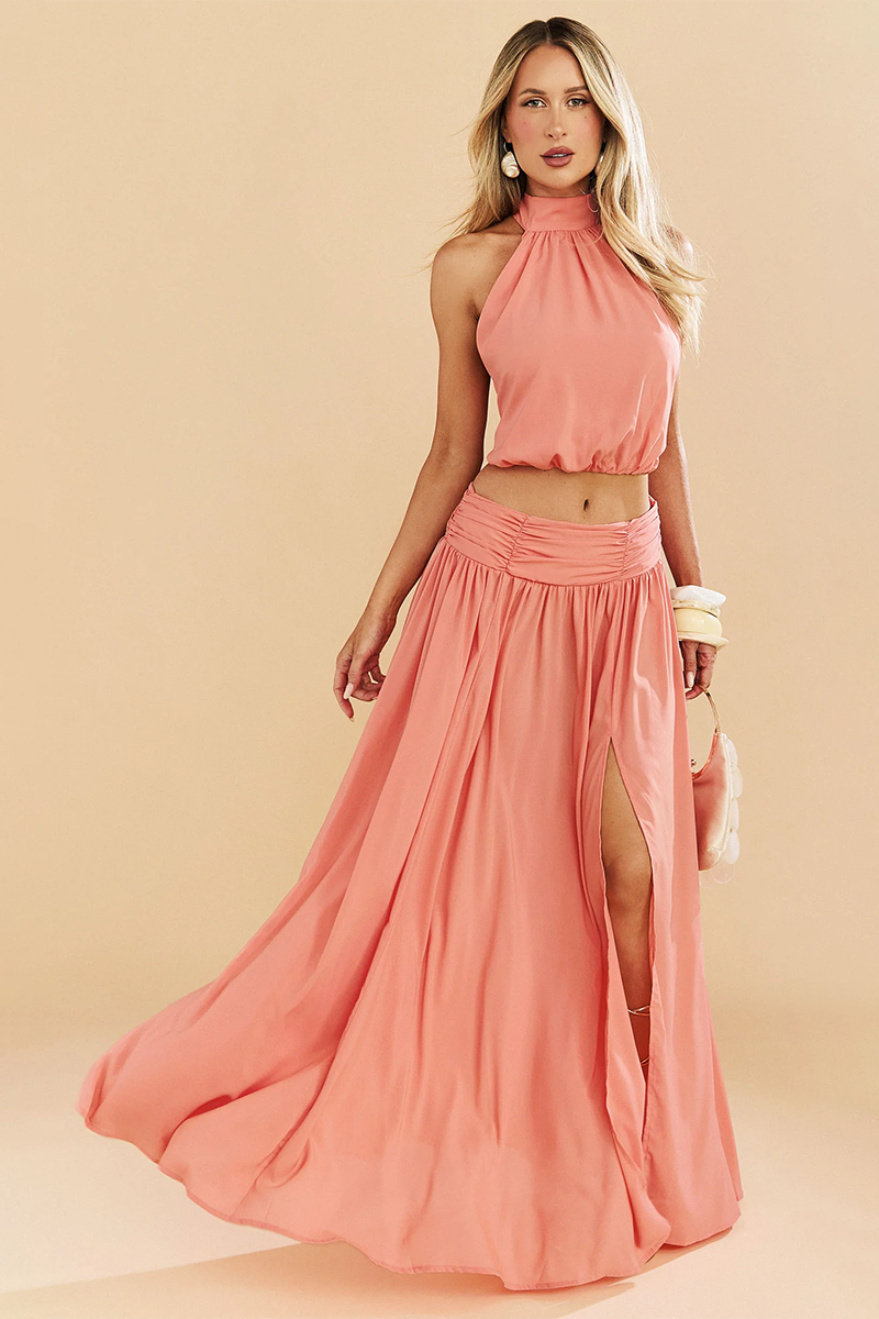 A woman wearing the coral-colored Showpo Melanie Set, featuring a halter-neck crop top and a matching maxi skirt with a high side slit.