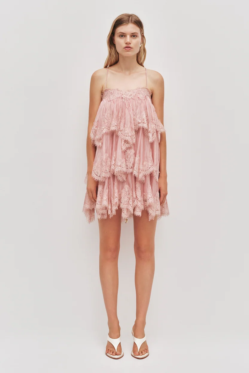 A model wears the fawn pink Aje Ophelia Lace Mini Dress, featuring a tiered design with spaghetti straps and white thong sandals.