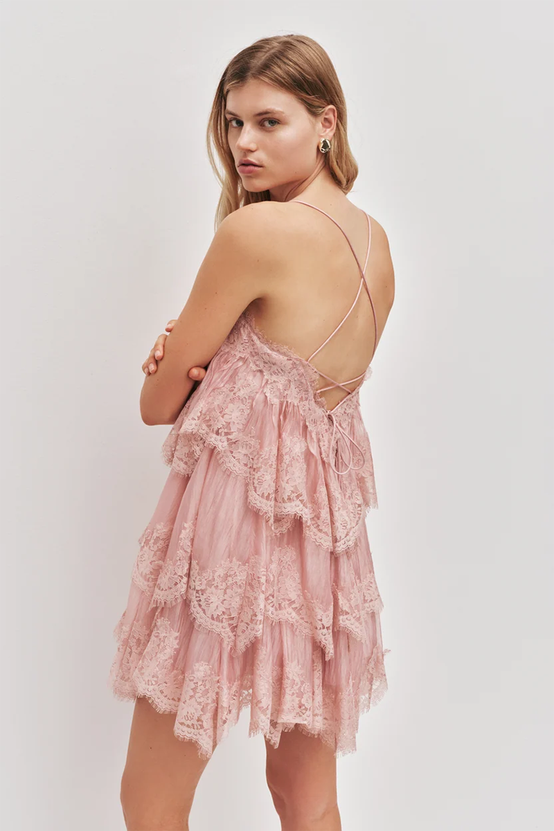 A woman wears the Aje Ophelia Lace Mini Dress in fawn pink, a tiered lace design featuring cross-back straps.