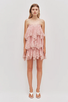 A model wears the fawn pink Aje Ophelia Lace Mini Dress, featuring a tiered design with spaghetti straps and white thong sandals.