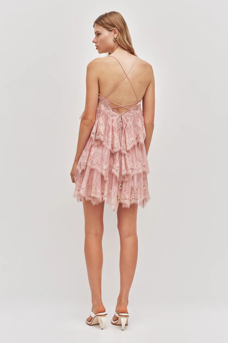 The Aje Ophelia Lace Mini Dress is a fawn pink lace dress with a tiered ruffled skirt and thin cross-back straps.