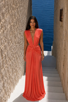 A woman wearing the Yasmina Gown by Abyss By Abby—a sleeveless, deep v-neck orange elegant maxi dress—stands on white stairs between stone walls, with the sea visible in the background.