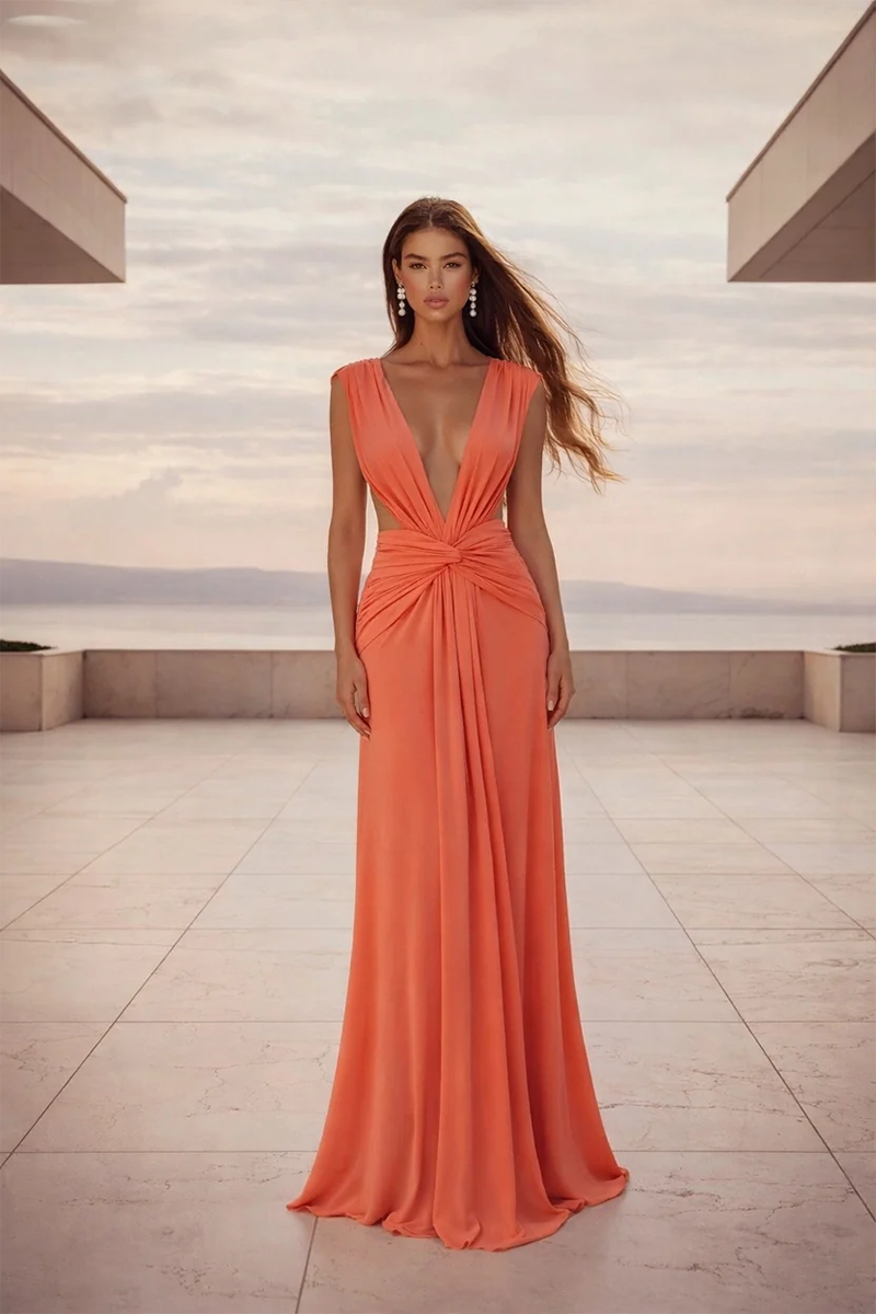 Wearing the Yasmina Gown by Abyss By Abby—a flowing, deep orange, figure-flattering dress with a plunging neckline—a woman stands on a tiled terrace, modern buildings and a sunset sky in the background.