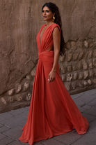 Wearing the Yasmina Gown by Abyss By Abby—a sleeveless, flowing orange maxi dress with a gathered waist—she stands on a stone path beside a textured brown wall, gazing ahead with long, wavy hair and gold earrings.