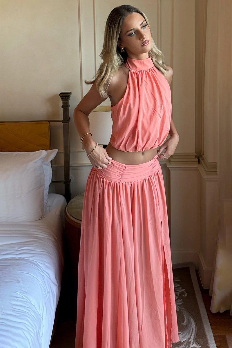 A woman wears the coral-colored Showpo Melanie Set, featuring a halter-neck crop top and a high-waisted maxi skirt with a side slit.