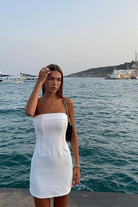A woman wearing the With Jean Chloe Corset Dress stands by the sea with boats and a lighthouse in the background on a clear day.